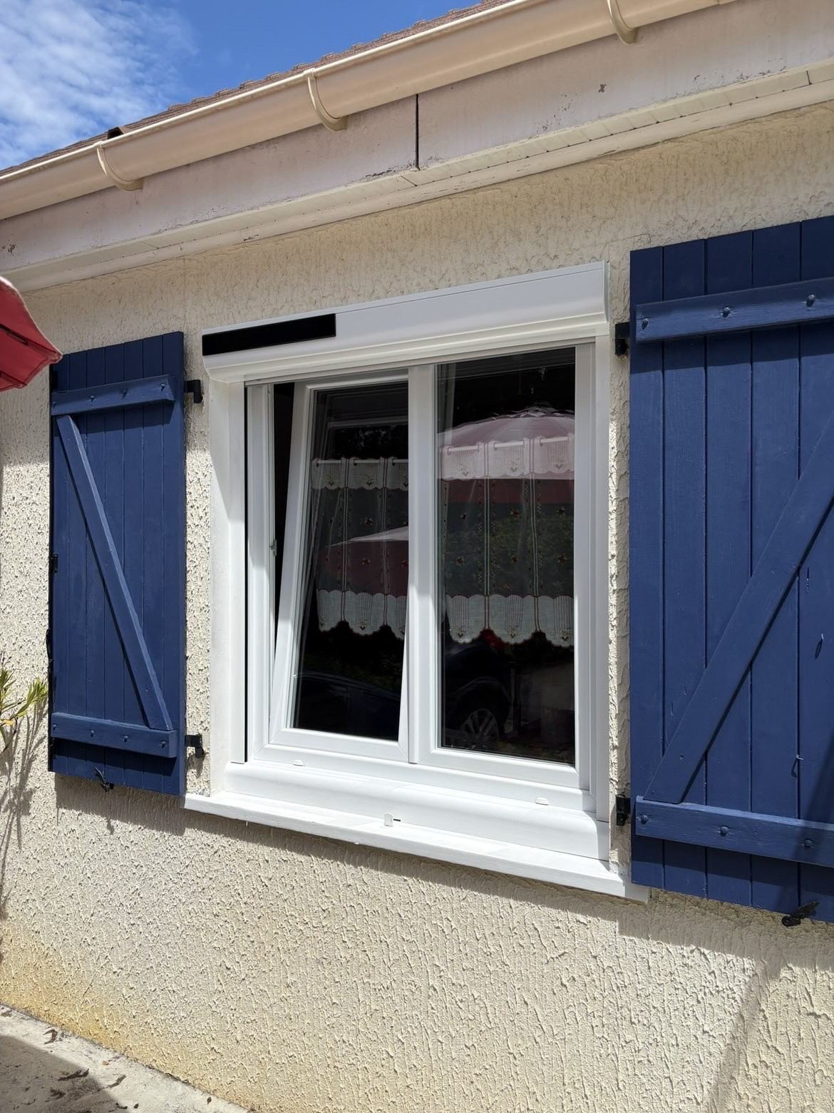 Fenêtre PVC blanche neuve avec volet roulant à énergie solaire, posée entre des volets battants bleus